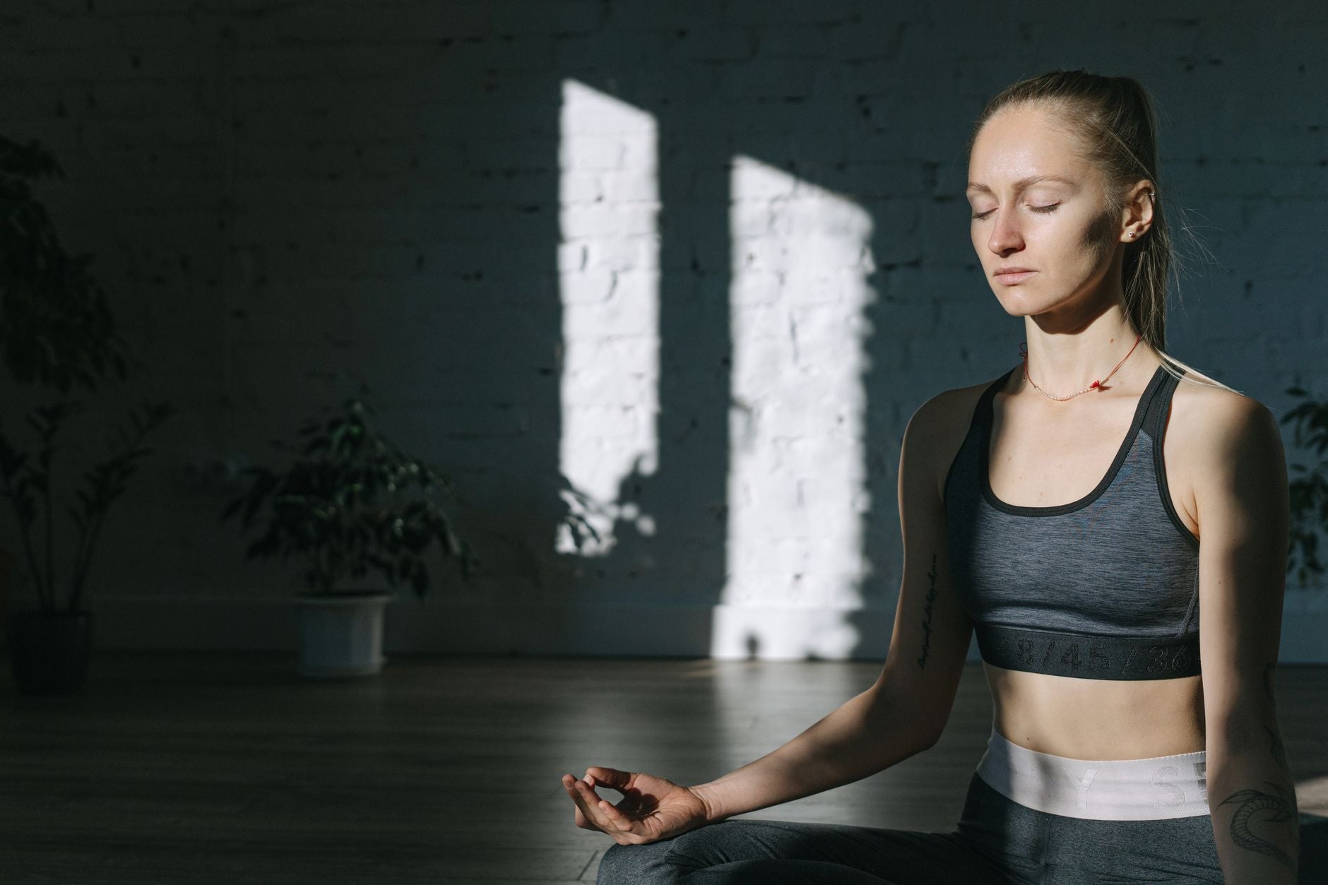 A woman in a calm seated meditation posture by a window — eyes closed, hands resting, breath visibly steady